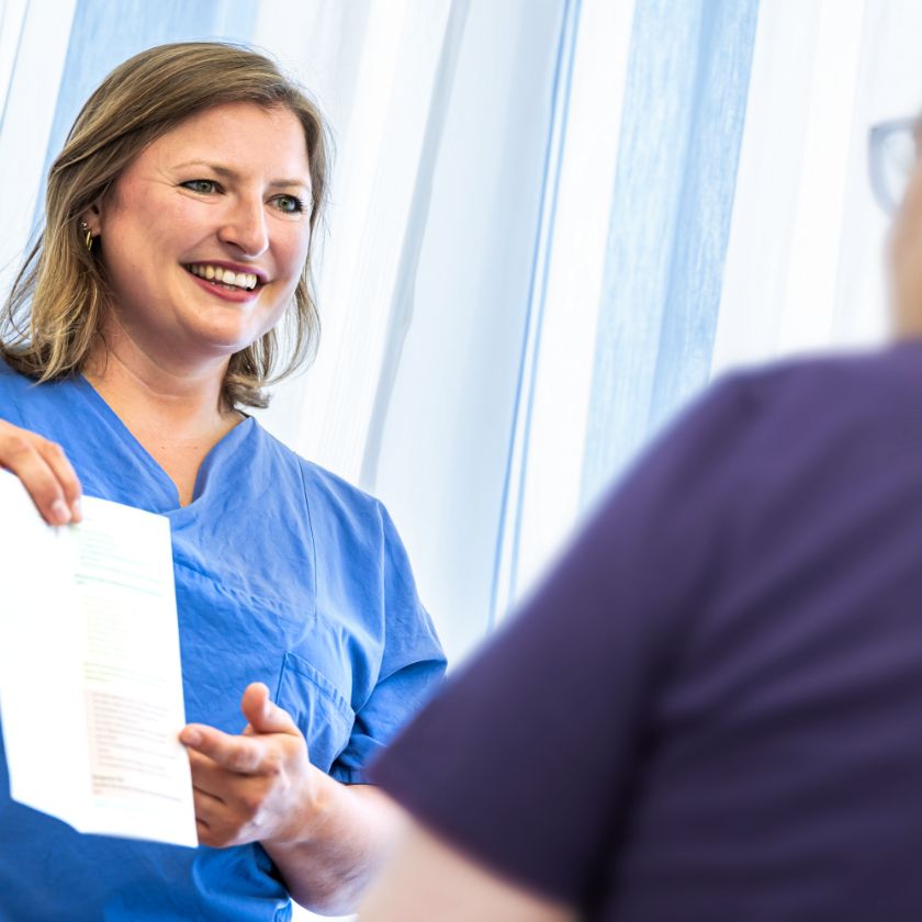 Pflegeexpertin Lena Jung zeigt einem Patienten ein Informationsblatt und lächelt. Der Patient ist unscharf von hinten zu sehen.