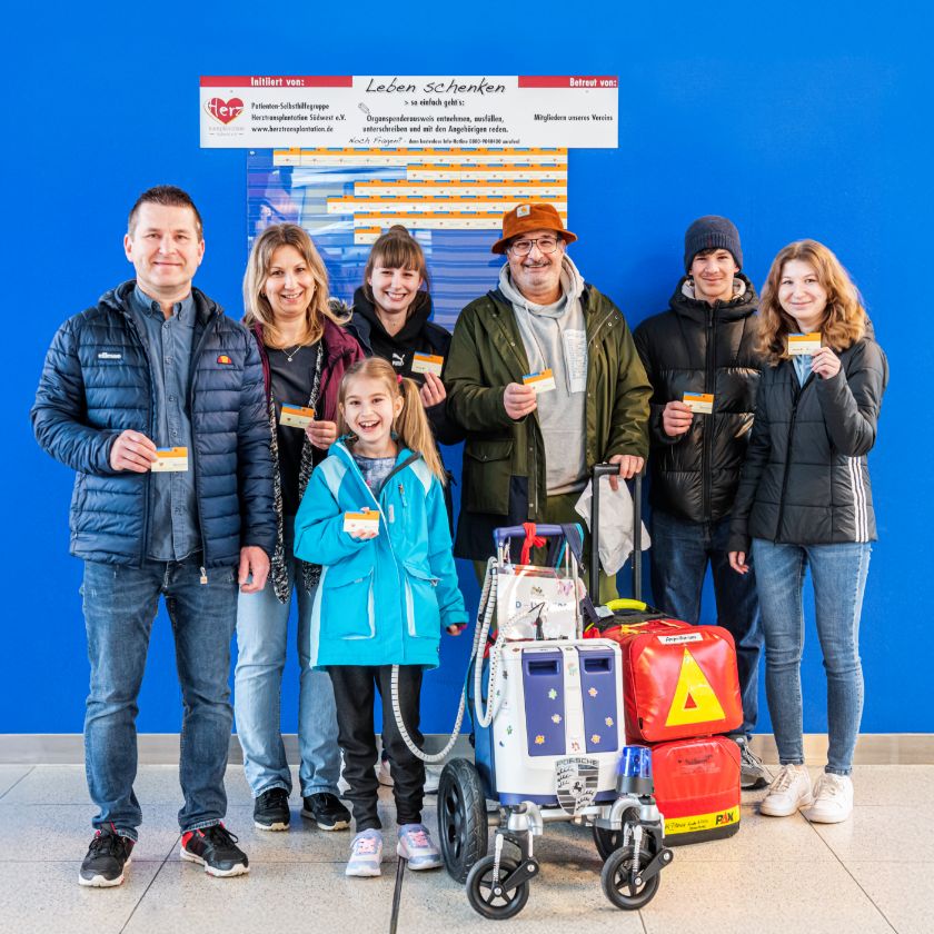 Gruppenfoto von sechs Erwachsenen und Lara, der Patientin aus der Kinderherzchirurgie. Neben ihr steht ein Wagen mit dem Berlin Heart. Alle blicken in die Kamera und halten einen Organspendeausweis in der Hand.