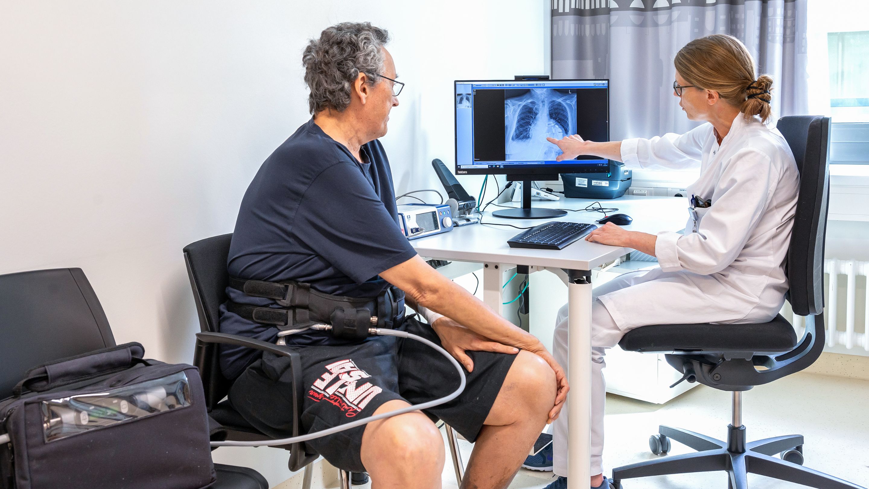 Prof. Dr. Meyer und ein Patient mit Kunstherz blicken gemeinsam auf einen Monitor, der ein Röntgenbild des Oberkörpers mit dem Kunstherz zeigt. Neben dem Patienten steht eine schwarze Tasche auf einem Stuhl, aus der ein weißer Schlauch zum Körper führt.