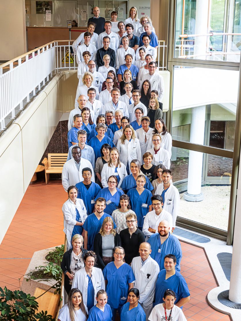 Gruppenfoto: Zahlreiche Personen in weißer und blauer medizinischer Dienstkleidung stehen lächelnd auf einer Treppe. Die Aufnahme wurde aus erhöhter Perspektive gemacht.
