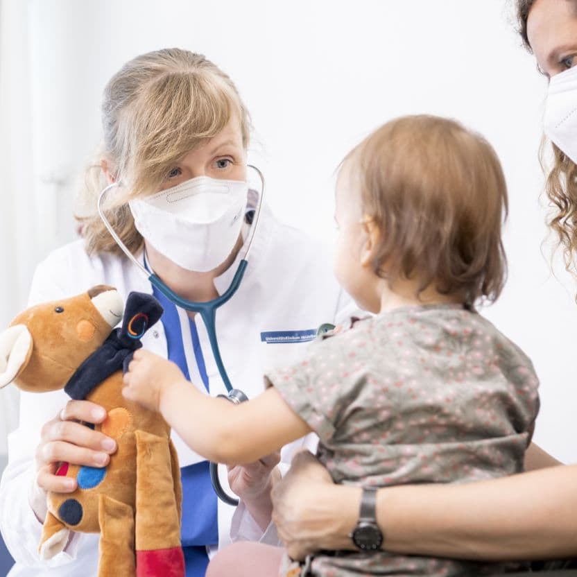 Eine Ärztin mit Stethoskop und Mundschutz sitzt einem Kleinkind gegenüber. Sie hält ein Kuscheltier-Reh in der Hand, nach dem das Kind greift. Das Kind ist unscharf von hinten zu sehen.