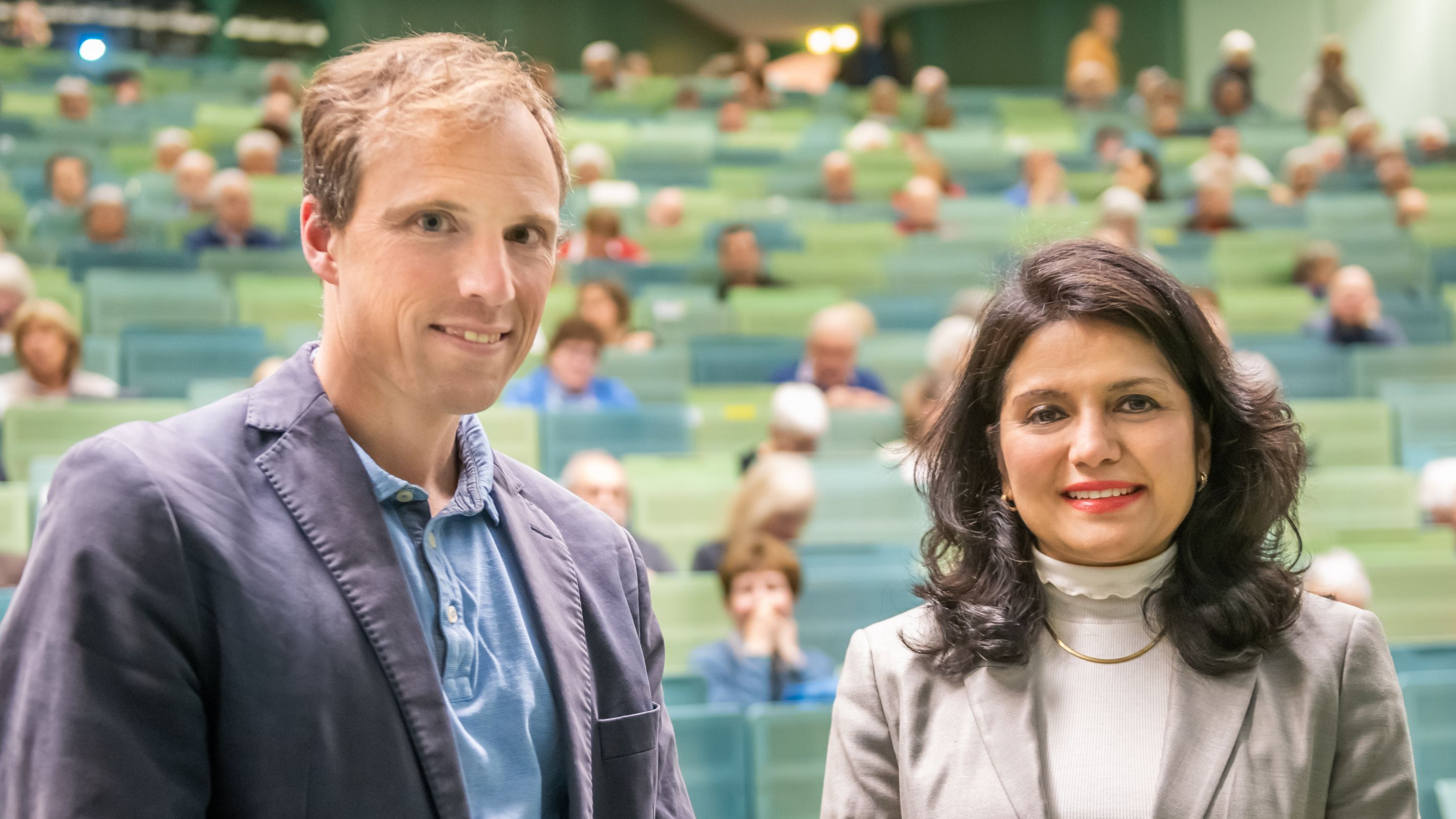 Prof. Dr. Jonas Tesarz und Prof. Dr. Rohini Kuner lächeln in die Kamera. Im Hintergrund sind unscharf die Reihen eines Hörsaals zu erkennen.