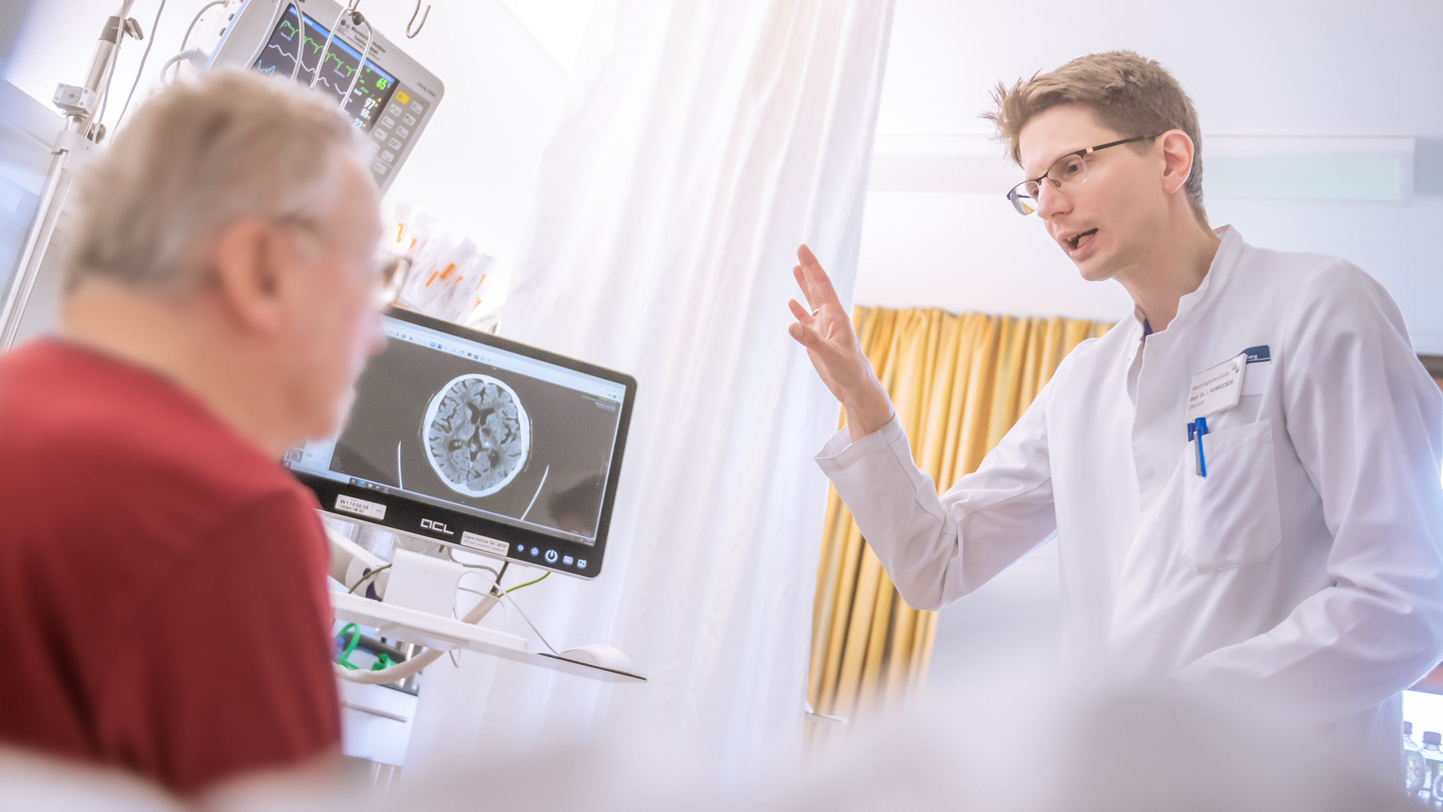 Eine medizinische Fachkraft im weißen Kittel spricht gestikulierend mit einem Patienten. Im Hintergrund zeigt ein Monitor ein MRT-Bild eines Kopfes, der Patient ist unscharf von hinten zu sehen.