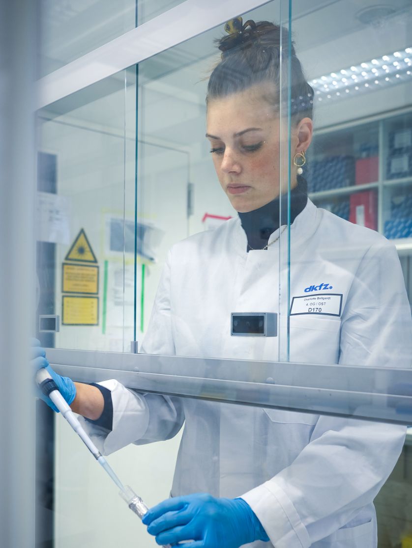 Eine Person in Laborkleidung arbeitet mit einer Pipette hinter einer Glasschutzscheibe.