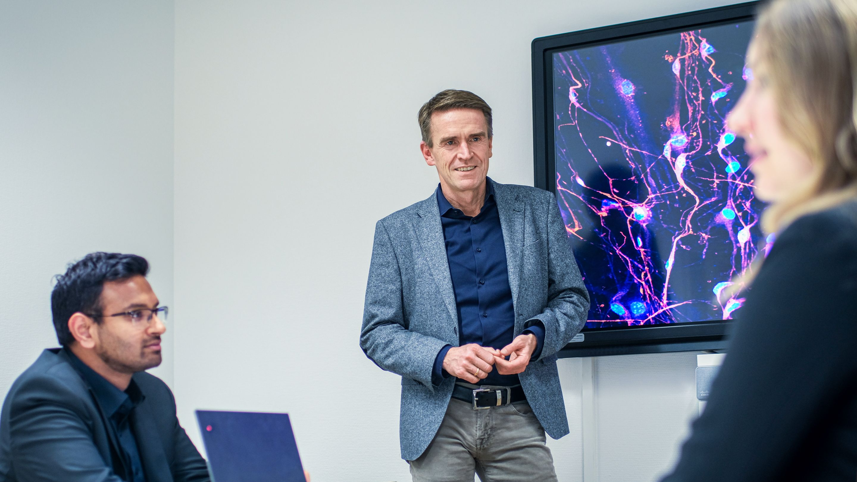 Prof. Dr. Wolfgang Wick mit zwei weiteren Personen in einem Besprechungsraum. Im Hintergrund zeigt ein großer Monitor leuchtende Linien und Punkte.