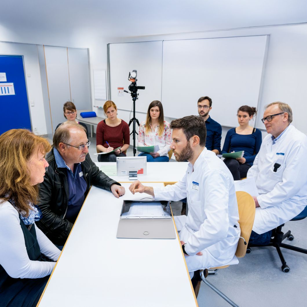Eine medizinische Fachkraft sitzt an einem Tisch zwei Personen gegenüber, neben ihr eine Kamera zur Aufzeichnung. Im Hintergrund weitere Beobachtende in Alltagskleidung sowie ein Arzt im Kittel.