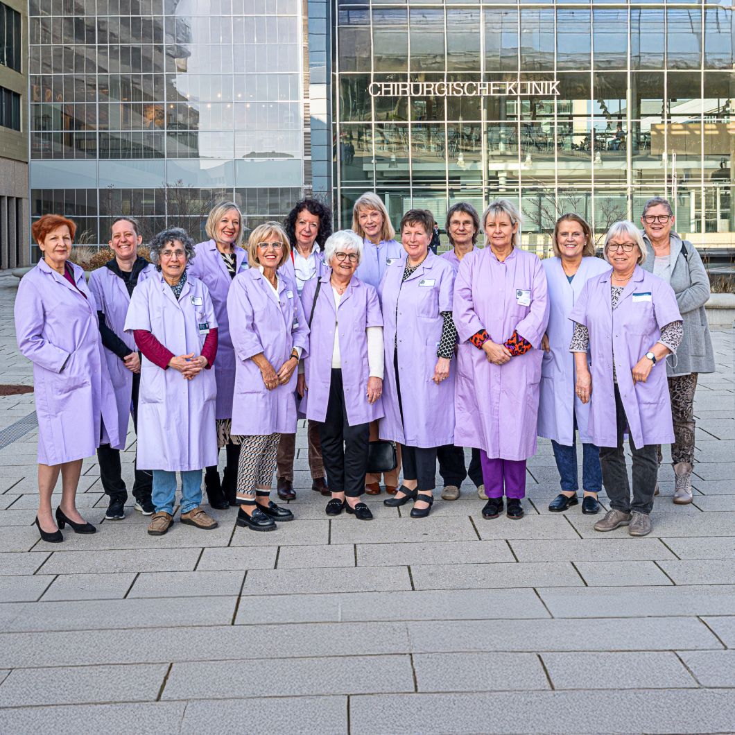 Gruppenfoto der ehrenamtlichen Lila Damen und Herren vor dem Eingang der Medizinischen und Chirurgischen Klinik. Die Personen stehen lächelnd nebeneinander in lila Dienstkleidung.