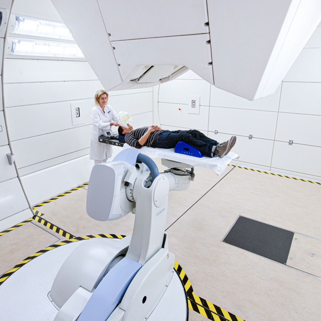 Ein Patient liegt in einem Behandlungsraum des Heidelberger Ionenstrahl-Therapiezentrums. Eine medizinische Fachkraft im weißen Kittel steht am Kopfende.