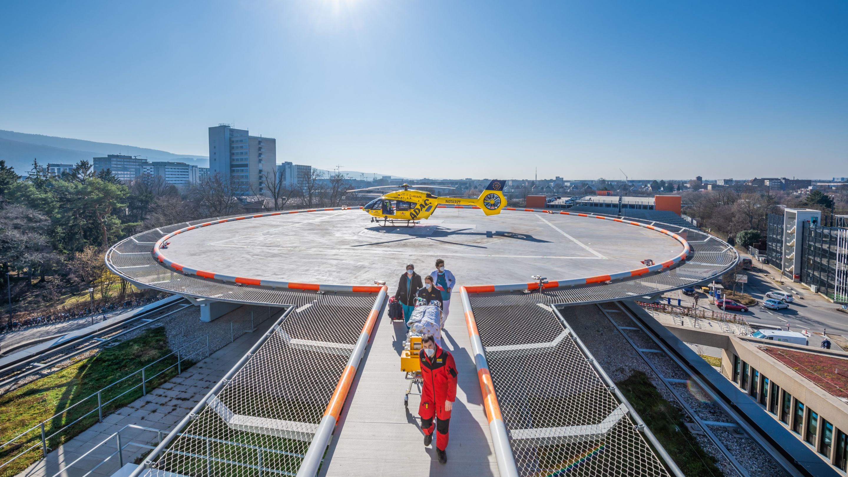 Ein gelber ADAC-Hubschrauber steht auf dem Landeplatz der Chirurgischen Klinik. Drei Personen begleiten eine Patientin oder einen Patienten auf einer Krankenliege über den Verbindungssteg zum Klinikeingang.