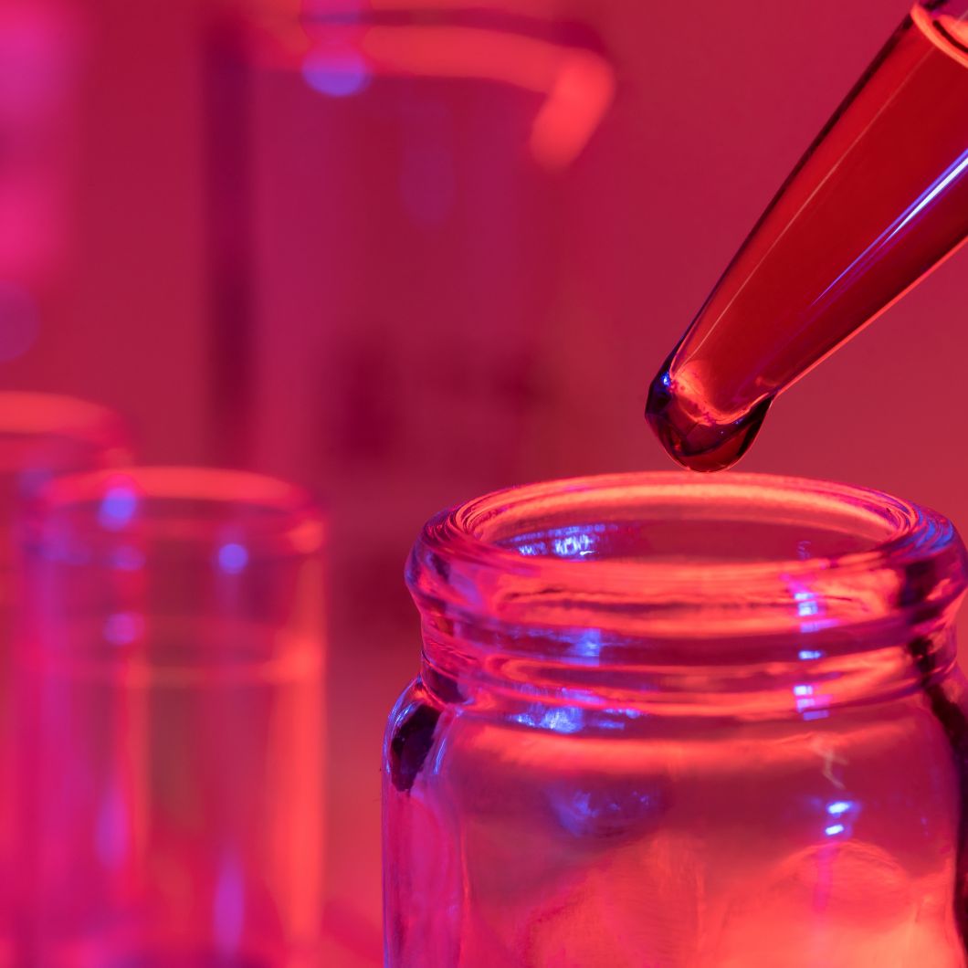 Symbolbild für Forschung: Eine Pipette über einem Glasrand, an der Spitze hängt ein Tropfen. Das Bild ist rot eingefärbt.