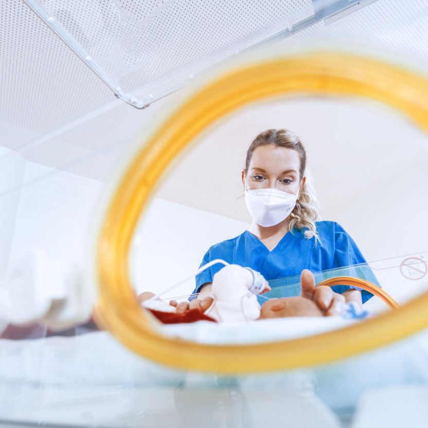 Eine Pflegekraft mit Mundschutz beugt sich über ein Frühchen im Inkubator und legt ihre Hände behutsam an Kopf und Bauch des Kindes. Der Blick fällt durch die seitliche Öffnung des Brutkastens; das Baby ist unscharf zu erkennen.