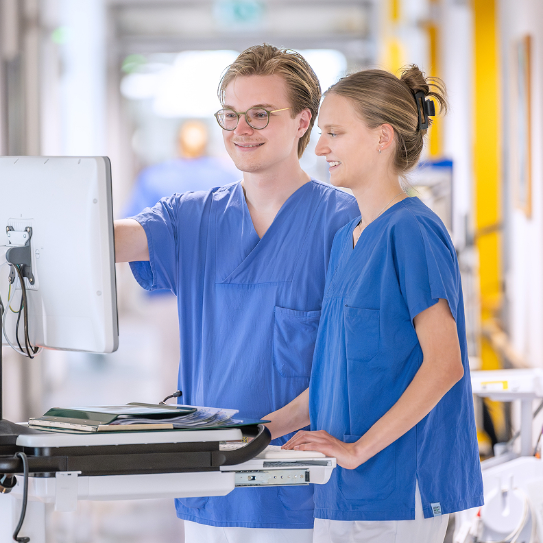 Zwei medizinische Fachkr&auml;fte in blauer Arbeitskleidung betrachten gemeinsam einen Computerbildschirm.