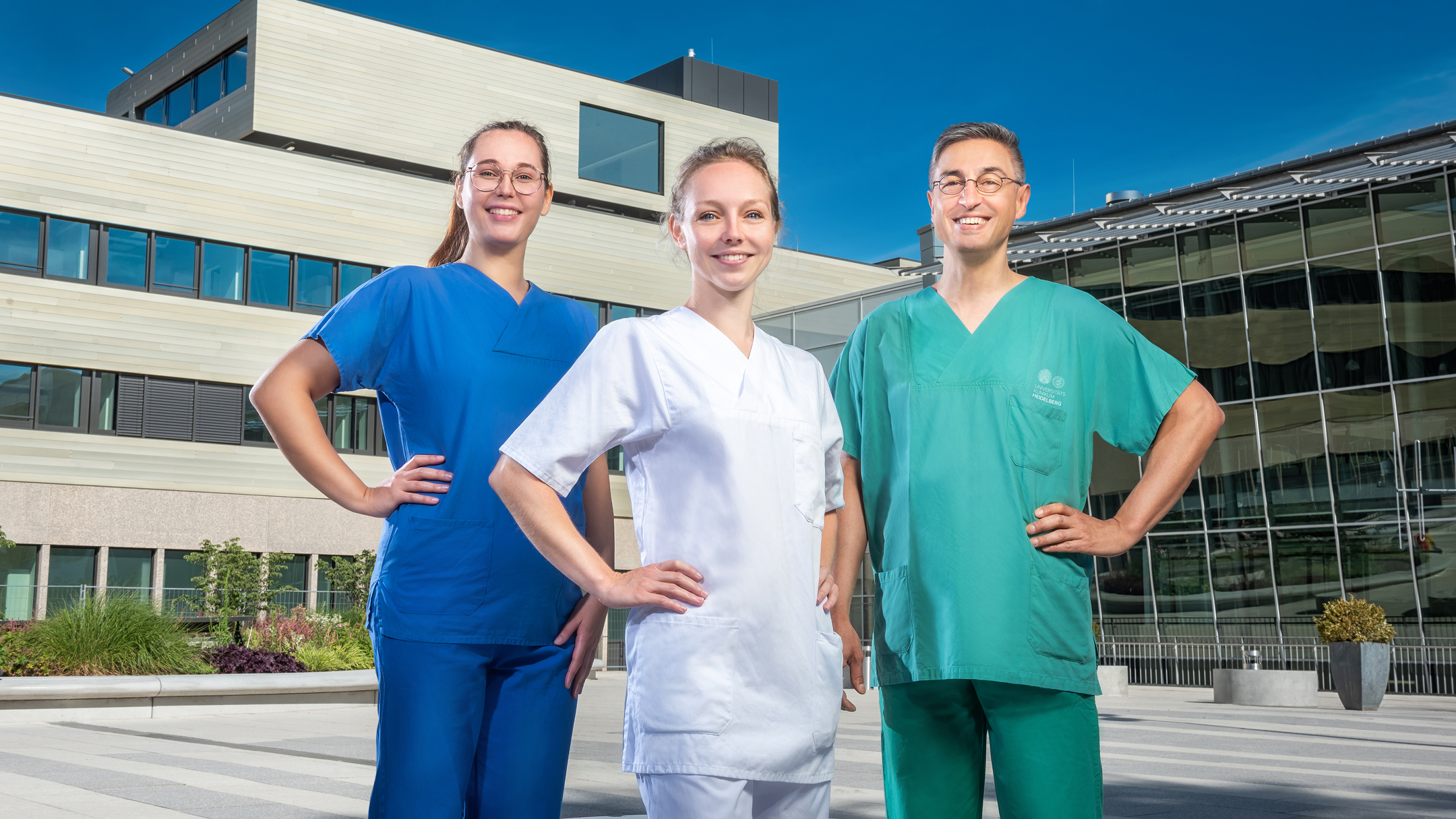 Drei medizinische Fachkr&auml;fte in blauer, wei&szlig;er und gr&uuml;ner Arbeitskleidung stehen vor einem Klinikgeb&auml;ude.