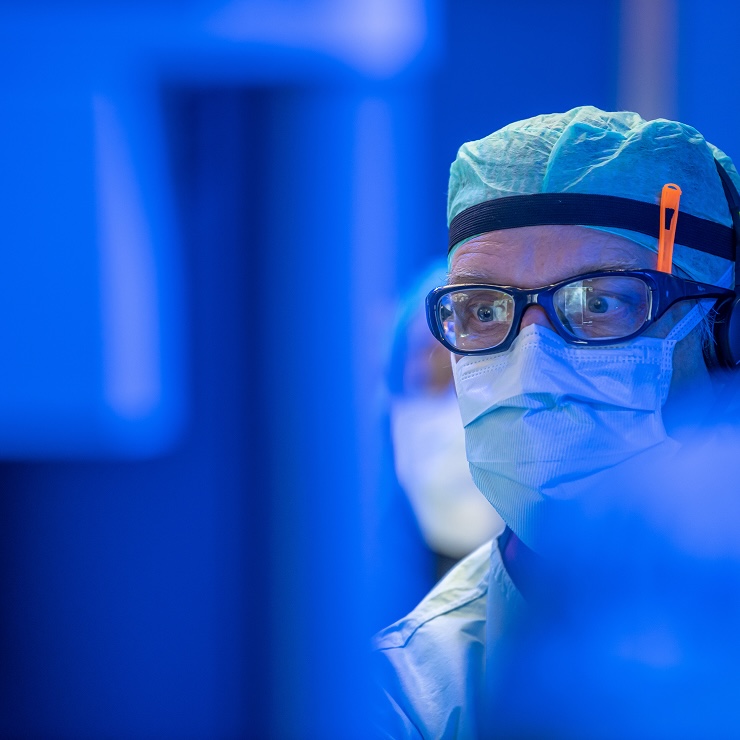 Eine medizinische Fachkraft mit Mundschutz, Schutzbrille und Haube in blau beleuchteter Umgebung.